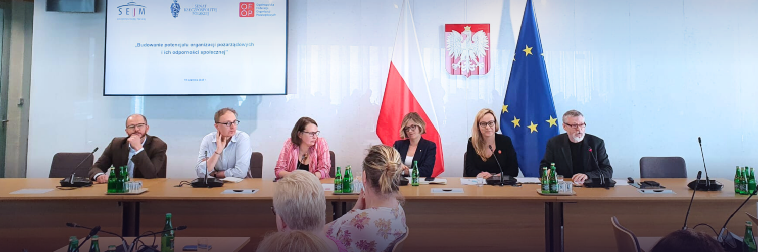 Sześć osób siedzi przy rzędzie stołów przodem do aparatu. Kontekst wyraźnie oficjalny. Na stołach stoją konferencyjne mikrofony. Za osobami pod ścianą stoją duże flagi, polska i Unii Europejskiej. Na ścianie godło Polski i ekran z prezentacją.