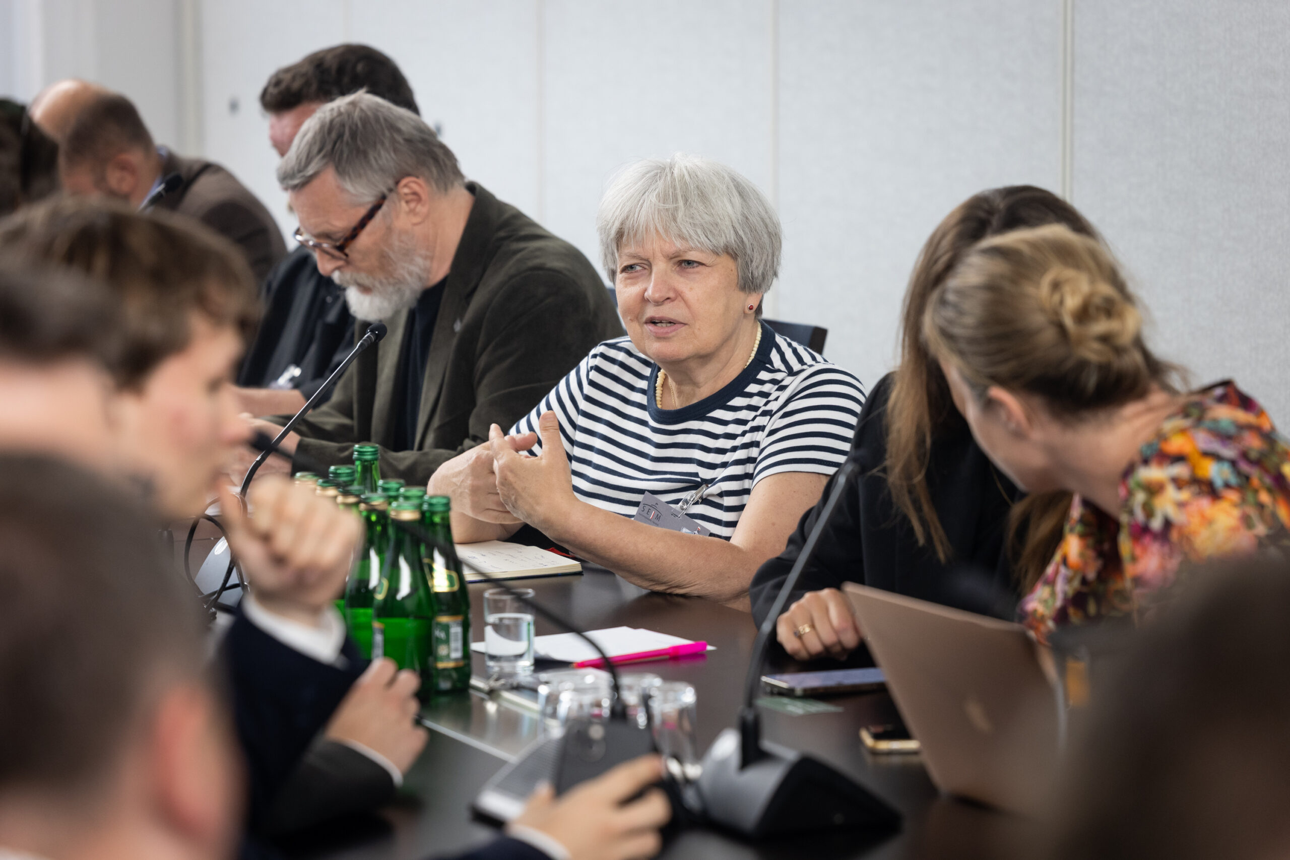 Zbliżenie na przemawiającą do mikrofonu osobę. Siedzi przy stole, z obu stron siedzą osoby z komputerami i telefonami.