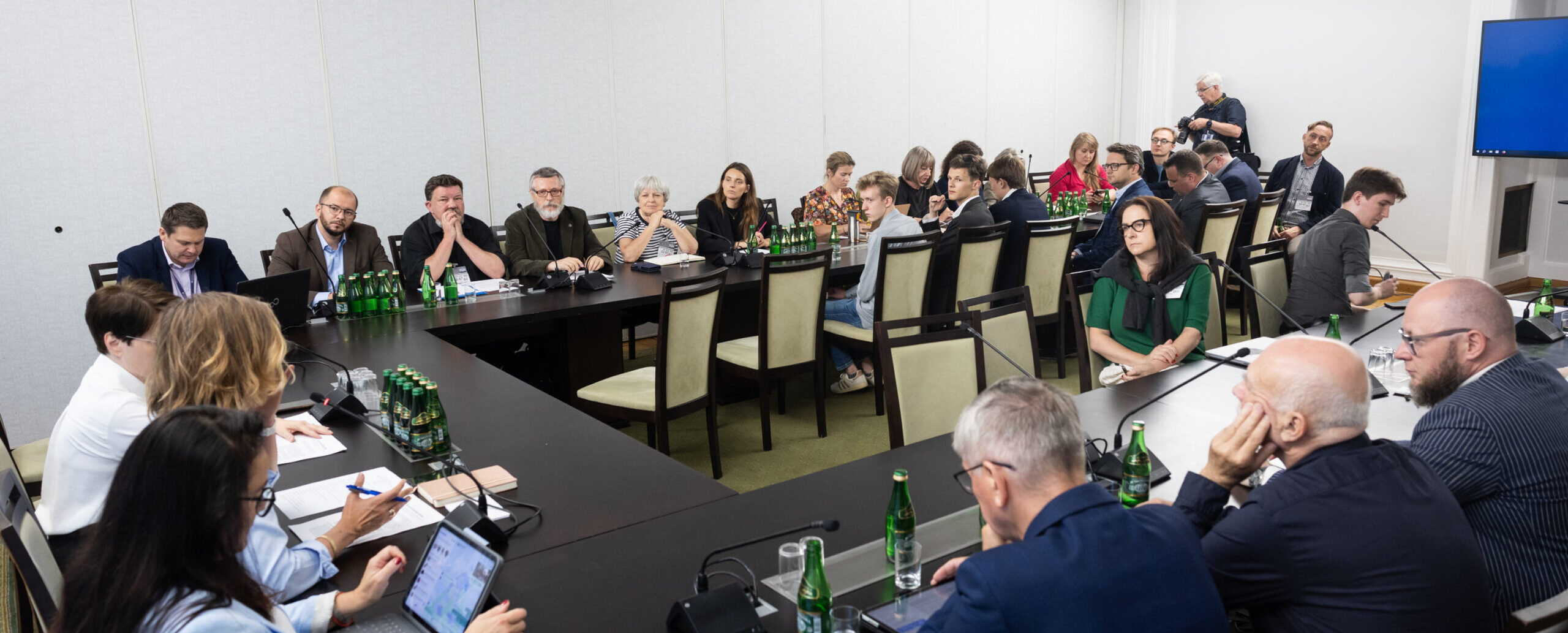Szerokie ujęcie na salę konferencyjną. Trzy stoły złączone w literę U. Na szczycie siedzą trzy osoby, prawdopodobnie prowadzące spotkanie. Przy wszystkich stołach siedzą osoby z komputerami i telefonami, każdy ma mikrofon konferencyjny ustawiony na blacie stołu. Sytuacja wygląda oficjalnie.